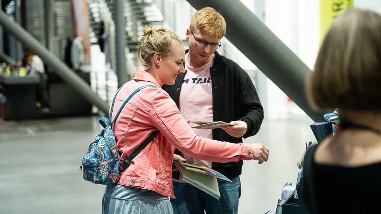Zwei Personen stehen im Foyer der Jahrhunderthalle Bochum und schauen sich einen Flyer der Ruhrtriennale an.