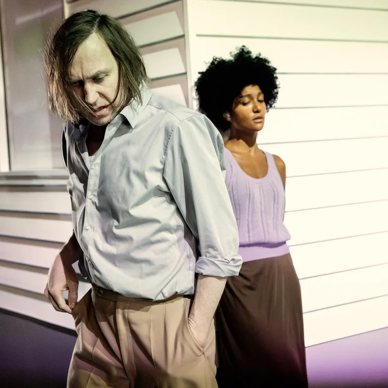 Larissa Sirah Herden mit verschränkten Armen steht ruhig im Hintergrund, während Lars Eidinger im Vordergrund mit geneigtem Blick vorbeigeht – die Szene wirkt angespannt.