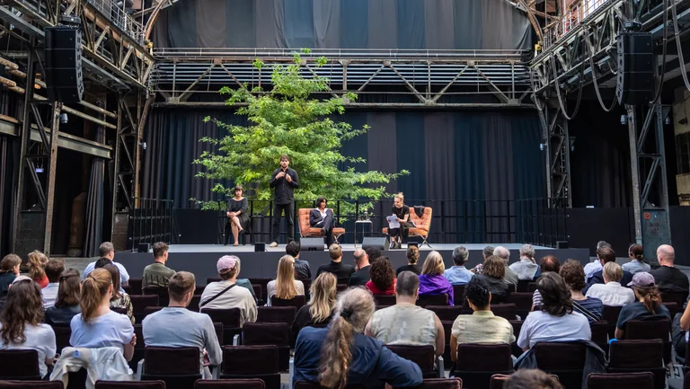 Diskussionsveranstaltung der Reihe Brave New Voices bei der Ruhrtriennale 2024 in der Jahrhunderthalle Bochum: Vier Personen sitzen bzw. stehen auf einer Bühne vor einem großen Baum, umgeben von industrieller Architektur. Im Vordergrund folgt ein Publikum aufmerksam der Gesprächsrunde.