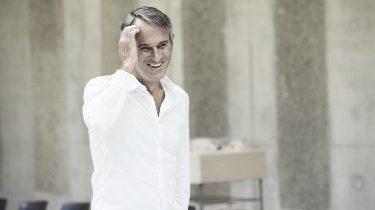 Ivo van Hove, Intendant der Ruhrtriennale 2024–2026, trägt ein weißes Hemd. Er greift sich mit der rechten Hand an die Stirn und lächelt.