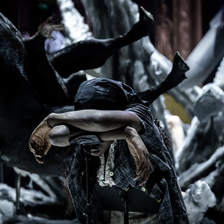 Eine Schauspielerin beugt sich erschöpft mit verschränkten Armen und gesenktem Kopf nach vorne. Hinter ihr ragen chaotisch geformte Kulissenteile und Skulpturen in Form von Körperteilen hervor. Die Atmosphäre ist bedrückend und düster.