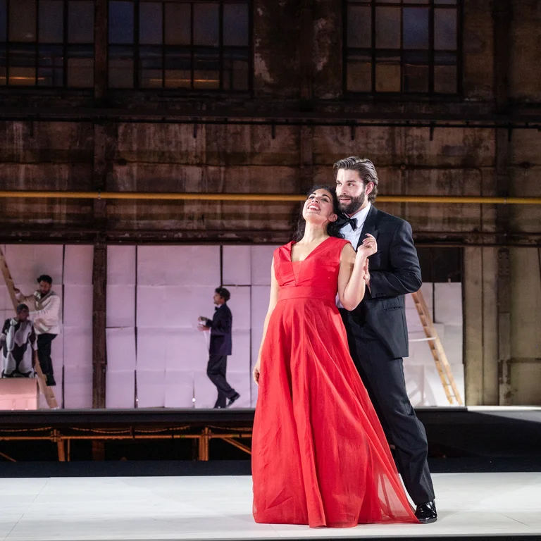 Eine Frau in Abendkleid und ein Mann in Anzug, nah beieinander stehend. Im Hintergrund weitere Personen, die weiße Papiere an die Wand hängen.