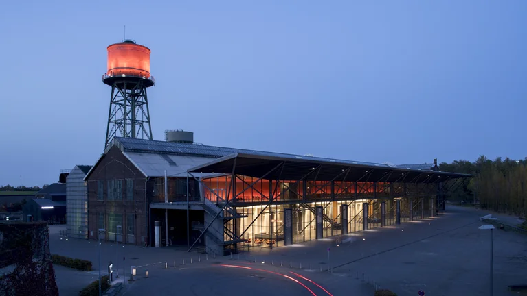 Die Jahrhunderthalle Bochum bei Nacht, beleuchtet durch ein markantes rotes Licht am Wasserturm und den Eingangsbereich, mit Spuren von Autolichtern im Vordergrund.