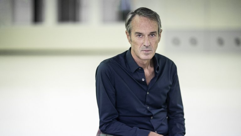 Ivo Van Hove, Intendant der Ruhrtriennale 2024–2026, sitzt auf einem karierten Sessel. Er trägt ein dunkelblaues Hemd und blickt direkt in die Kamera.
