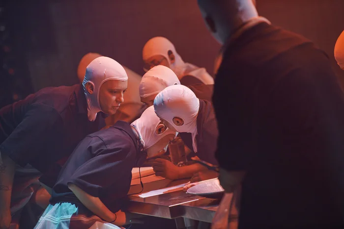 Several performers wearing white head coverings lean intently over a table, deeply engaged in a shared action. The red stage lighting enhances the intense and mysterious atmosphere.