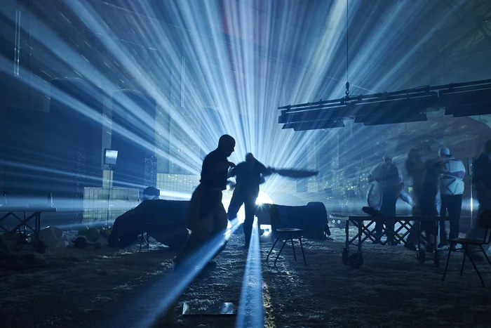 Silhouettes of performers move in front of a radiant fan of blue light spreading across the space. The scene feels dynamic and intense, as if marking a climax of the performance.