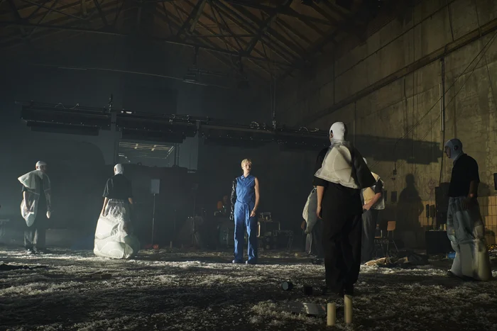 Several performers stand in a dimly lit space, with one person in a blue jumpsuit at the center. The scene feels theatrical and mysterious, emphasized by unusual costumes and subdued lighting.