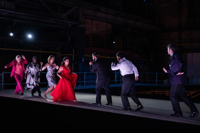 Mehrere Performer:innen bewegen sich dynamisch auf einer hellen, erhöhten Bühne in einer abgedunkelten Industriehalle. Links im Bild tragen Helena Rasker ein pinkfarbenes Kostüm, Claron McFadden ein schwarz-weißes Gewand, Nina van Essen ein silbernes Paillettenkleid und Sophia Burgos ein rotes Abendkleid. Ihnen gegenüber stehen männliche Performer in eleganter Abendgarderobe.