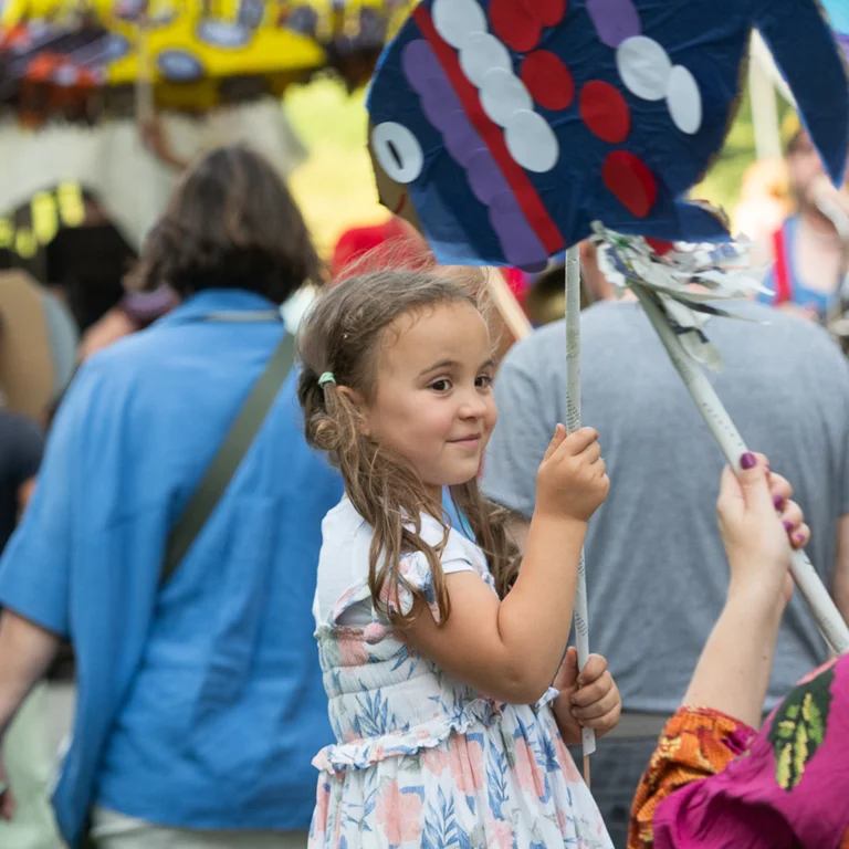 A joyful girl with long brown hair holds a colorful, handmade paper fish on a stick. She wears a summery floral dress and smiles while looking to the side. In the background, many people dressed in colorful clothing participate in a festive parade or procession.