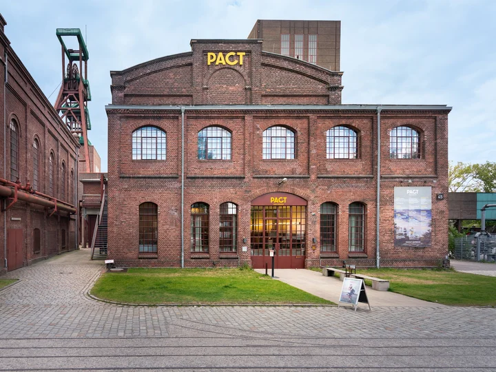 Außenansicht des PACT Zollverein-Gebäudes in Essen mit roten Backsteinwänden und großen Fenstern, im Hintergrund der Förderturm.