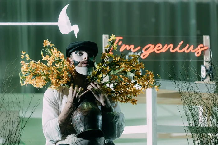 A person with a white mask holds a vase of yellow flowers, with the slogan "I'm genius" in neon lettering in the background.