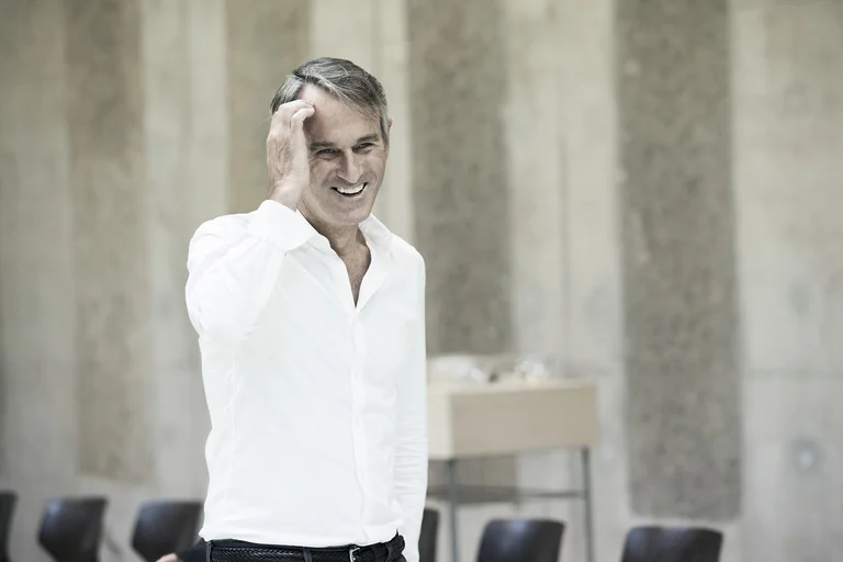 Ivo van Hove, Intendant der Ruhrtriennale 2024–2026, trägt ein weißes Hemd. Er greift sich mit der rechten Hand an die Stirn und lächelt.