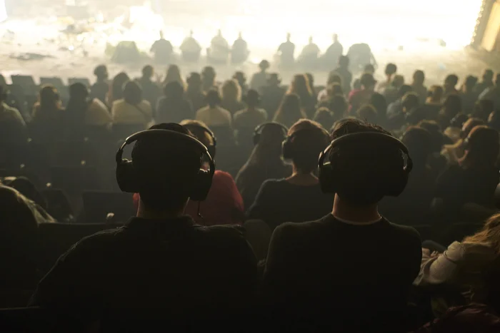 View from the back of the auditorium toward a densely seated audience. All spectators are wearing large headphones, which are part of the performance experience. The stage area in front fades into bright light.