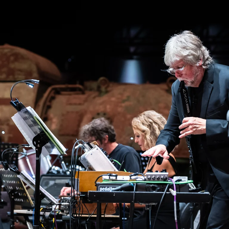 Ein Musiker des Will Gregory Moog Ensembles steht im Vordergrund und spielt konzentriert an einem analogen Synthesizer. Im Hintergrund sind weitere Musiker an ihren Instrumenten zu sehen, während die rostigen Turbinen der Industriehalle einen eindrucksvollen Kontrast zur modernen elektronischen Ausrüstung bilden.