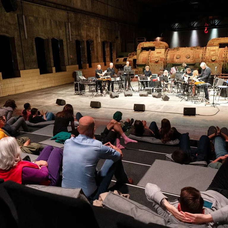Musicians perform on stage while the audience sits and lies relaxed in front.
