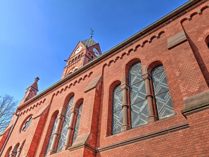 Eine Nahaufnahme der roten Backsteinfassade der Evangelischen Kirche am Katernberger Markt mit verzierten Rundbogenfenstern, dem hohen Kirchturm und einer blau schimmernden Glasstruktur in den Fenstern.