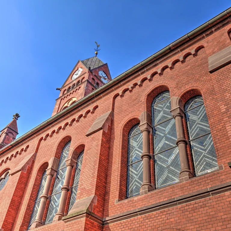 Eine Nahaufnahme der roten Backsteinfassade der Evangelischen Kirche am Katernberger Markt mit verzierten Rundbogenfenstern, dem hohen Kirchturm und einer blau schimmernden Glasstruktur in den Fenstern.