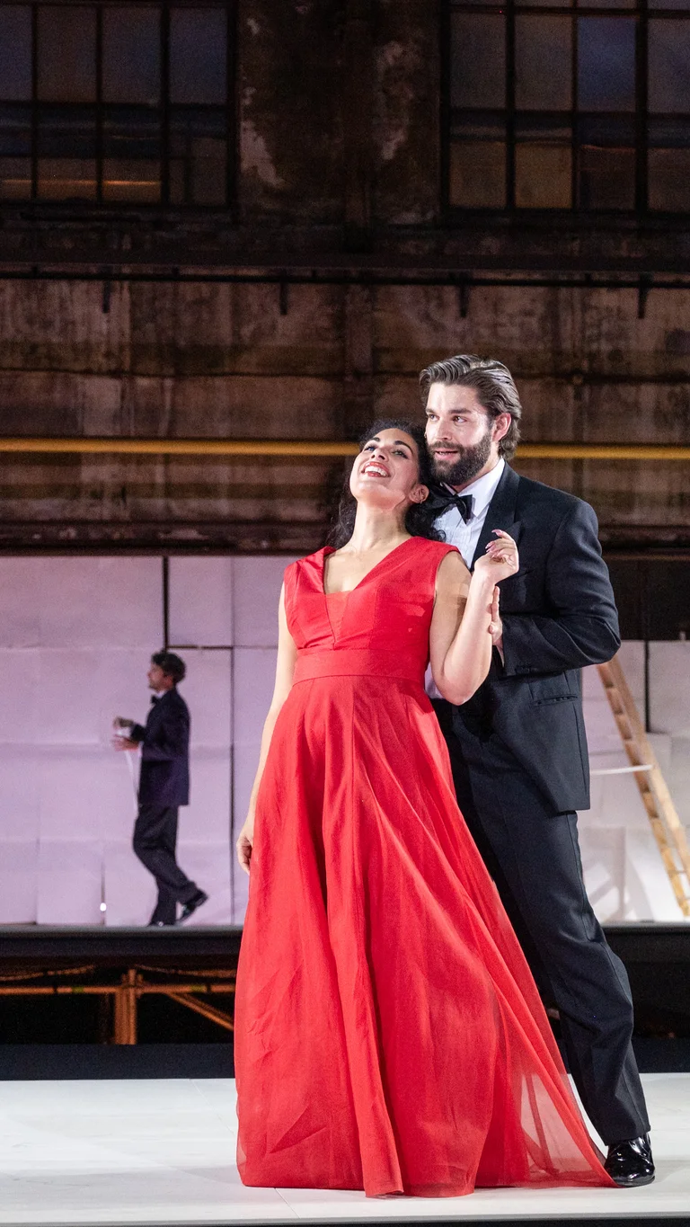 Eine Frau in Abendkleid und ein Mann in Anzug, nah beieinander stehend. Im Hintergrund weitere Personen, die weiße Papiere an die Wand hängen.