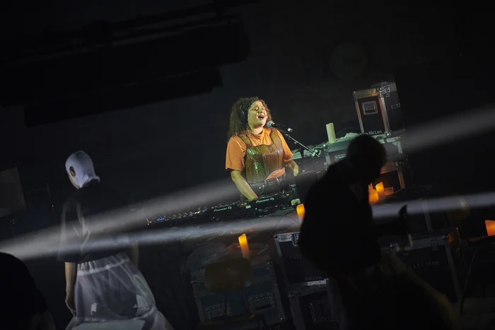 Gîn Bali performs passionately at a mixing desk, surrounded by candles and beams of light. In the foreground, shadowy figures of other performers contribute to the mysterious atmosphere.