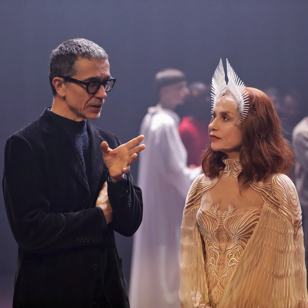 Romeo Castellucci talks to Isabelle Huppert, who is wearing a golden dress and a crown-like headdress. In the background we see people in conversation.