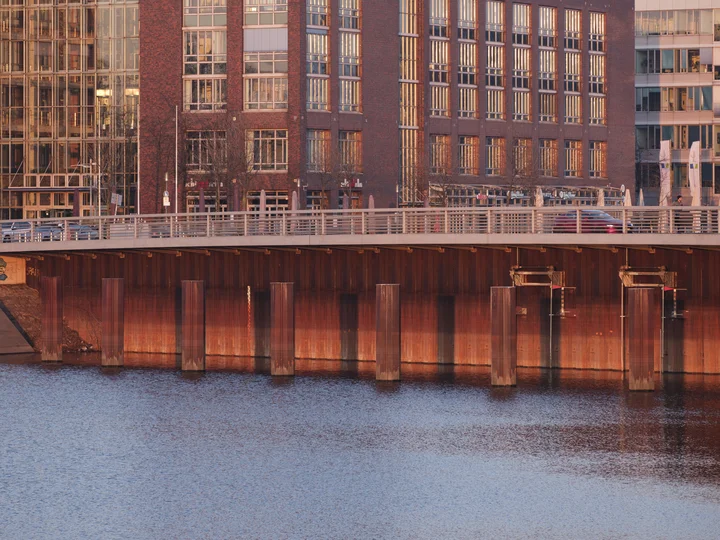 Eine Brücke über einem Fluss, deren rostrote Stahlpfeiler im Wasser reflektiert werden. Im Hintergrund spiegeln sich Backstein- und Glasgebäude im warmen Licht der untergehenden Sonne.