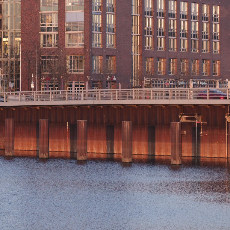 Eine Brücke über einem Fluss, deren rostrote Stahlpfeiler im Wasser reflektiert werden. Im Hintergrund spiegeln sich Backstein- und Glasgebäude im warmen Licht der untergehenden Sonne.