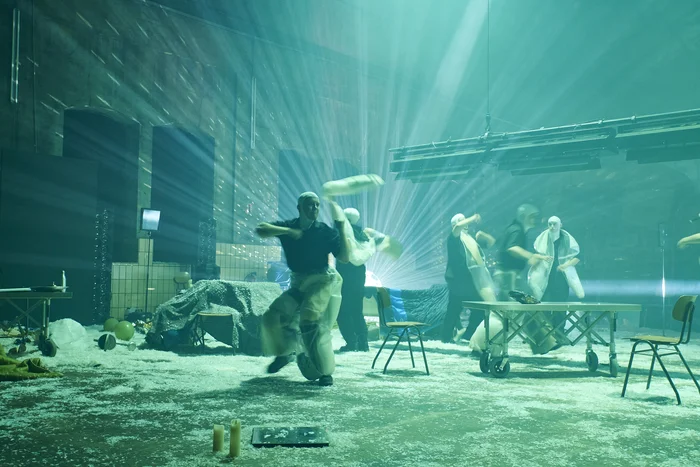Dance-like stage scene in a large hall bathed in green light and laser beams. Several performers wearing white hoods and transparent coverings move dynamically, some throwing or swinging pieces of fabric through the air. The floor is covered with white residue, scattered with chairs, tables, and various objects.