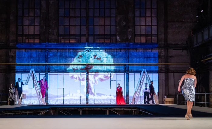 Mehrere Performer:innen stehen auf einer erhöhten Bühne vor der großen Fensterfassade einer ehemaligen Industriehalle. In der Bildmitte ist die Projektion einer atomaren Pilzwolke zu sehen, die eine dramatische Atmosphäre erzeugt. Rechts im Bild schreitet eine Frau in einem silbernen Paillettenkleid auf die Bühne zu, während andere Darsteller:innen zwischen Leitern und weißen Paneelen positioniert sind.