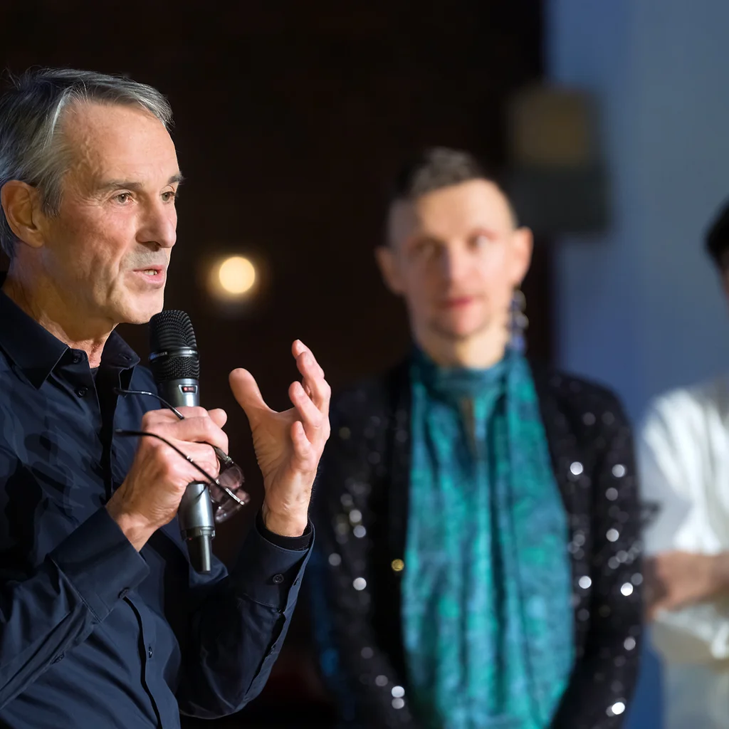 Ivo Van Hove speaks into a microphone, Tomasz Prasqual and Deniz Bolat stand in the background.