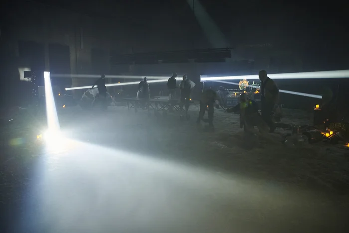 The scene depicts a dark performance with people in protective clothing, pierced by intense beams of light. The space feels mysterious and atmospheric, resembling a mix between a laboratory and a ritual.
