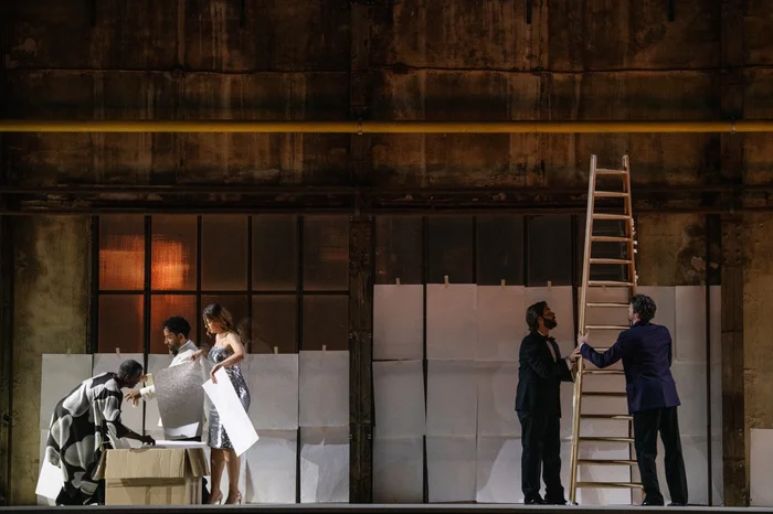 Fünf Performer:innen stehen auf einer erhöhten Bühne vor der Fensterfront einer alten Industriehalle. Links bückt sich Claron McFadden über eine Kiste, während Michael Wilmering und Nina van Essen weiße Papierbögen in den Händen halten. Rechts im Bild halten Alex Rosen und Steven van Linden eine hohe Holzleiter.