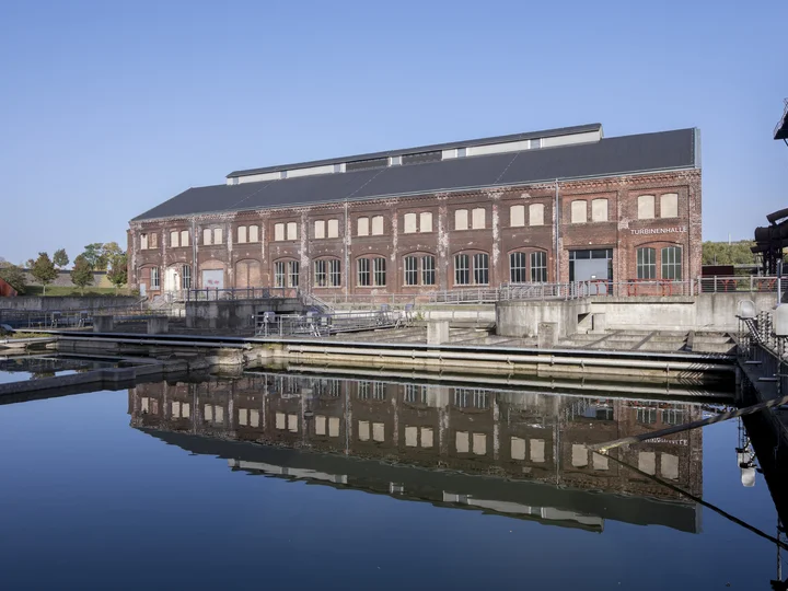 Die Turbinenhalle an der Jahrhunderthalle Bochum mit spiegelnder Wasserfläche im Vordergrund.