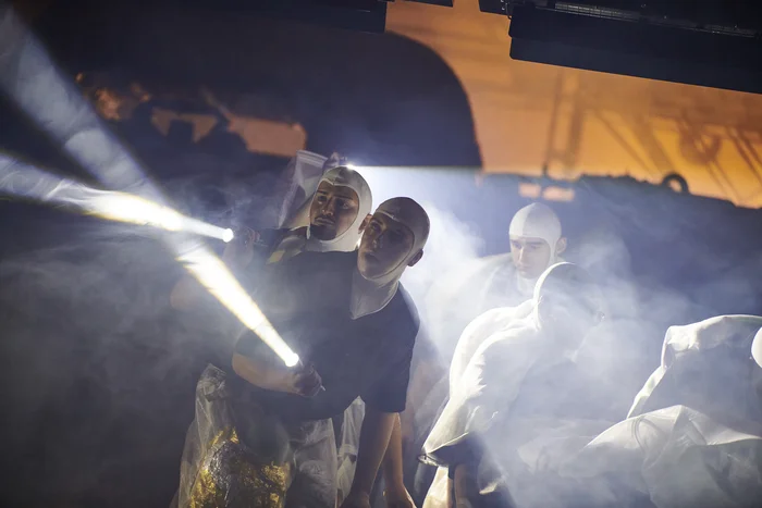 Several performers wearing white hoods and protective coverings move through dense fog. Holding flashlights, they shine intense beams of light into the darkness, cutting sharply through the haze. The scene feels tense and searching, almost like a rescue or exploration mission.