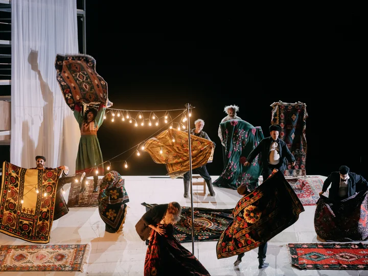 Eine Gruppe von Personen auf einer Bühne, die Teppiche hochhalten. Über ihnen leuchtet eine Lichterkette