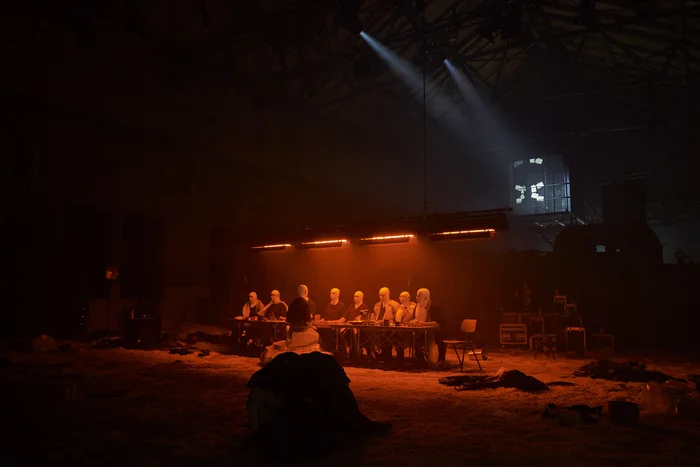 A group of performers wearing white head coverings sit at a long table bathed in red light. The dark space and dramatic spotlight from above enhance the mysterious, almost ritualistic atmosphere.