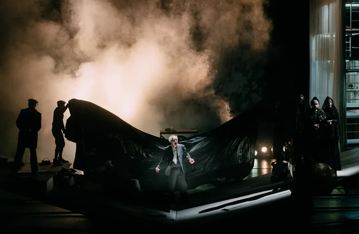 A man stands on a dark stage, fog rises behind him and a large black cloth blows.