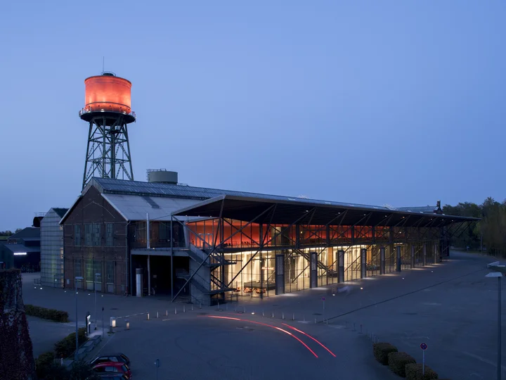 Die Jahrhunderthalle Bochum bei Nacht, beleuchtet durch ein markantes rotes Licht am Wasserturm und den Eingangsbereich, mit Spuren von Autolichtern im Vordergrund.