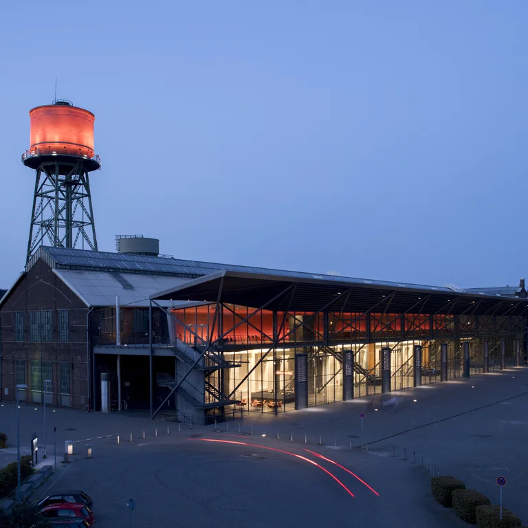 Die Jahrhunderthalle Bochum bei Nacht, beleuchtet durch ein markantes rotes Licht am Wasserturm und den Eingangsbereich, mit Spuren von Autolichtern im Vordergrund.