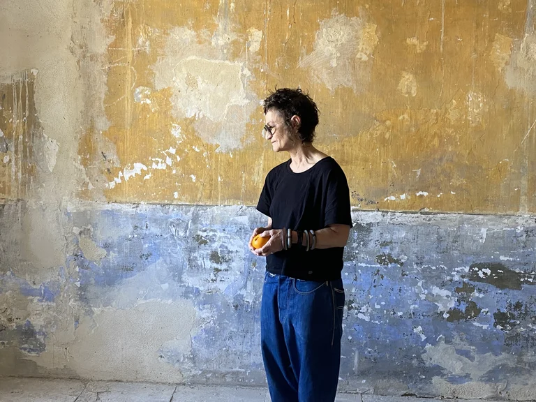 Robyn Orlin in schwarzem T-Shirt und blue Jeans vor einer verwitterten Wand mit ockerfarbenem und bläulich abgeplatztem Putz.